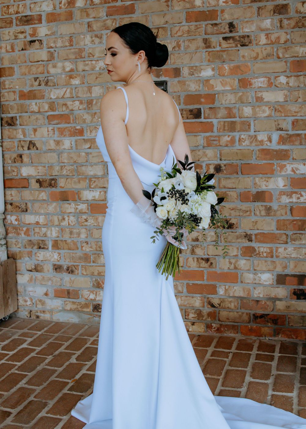 Bride in white dress holding bouquet poses against rustic brick wall.