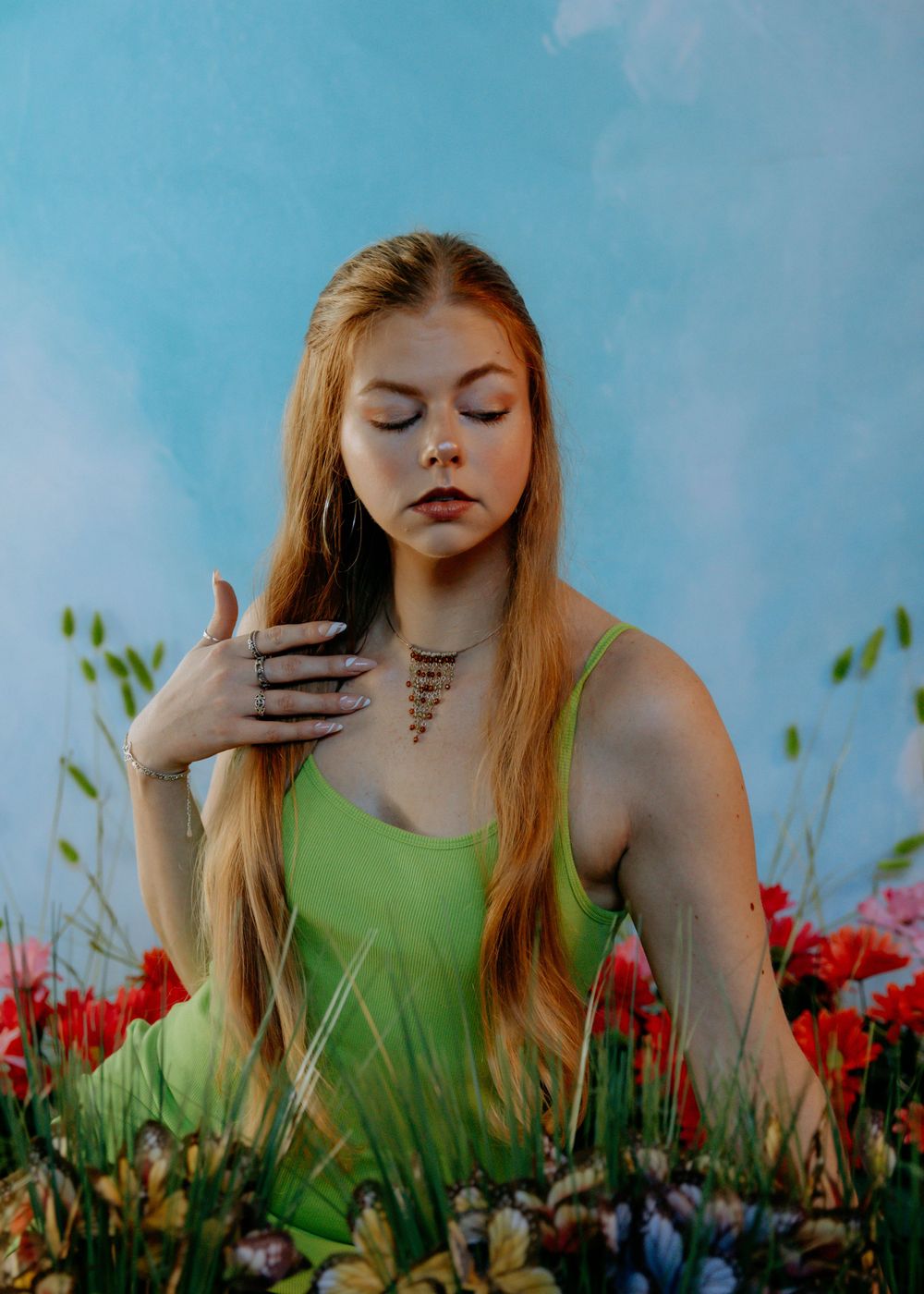 Artistic portrait in a flower field with green dress against vibrant blue sky and red blooms.