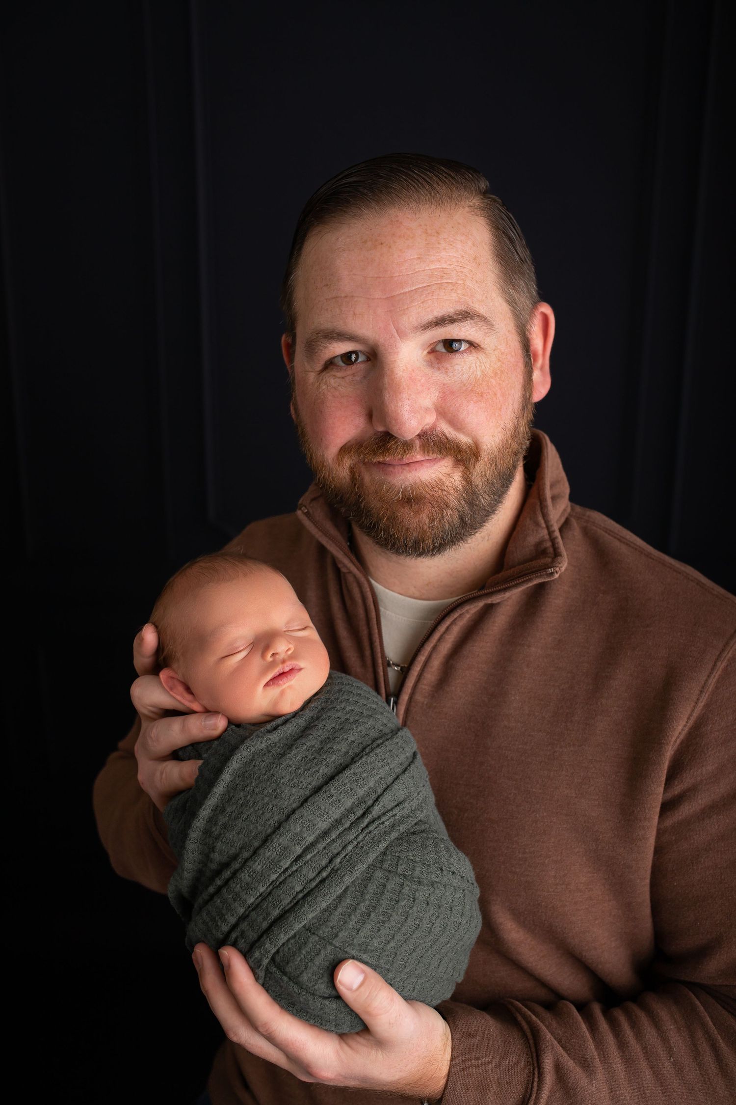 Portrait of parent and newborn baby wrapped in gray swaddle against dark background.