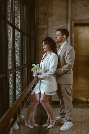 Couple in contrasting outfits pose by vintage windows with decorative details.