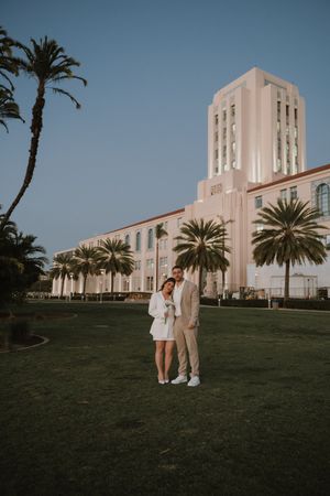 Couple poses in front of an Art Deco style building with palm trees.