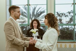 Elegant couple in white and beige outfits exchanging loving glances near large windows.