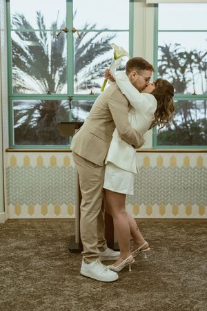 Romantic couple in formal wear dancing near windows with palm trees visible outside.