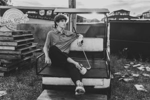 A vintage black and white photo showing someone lounging on the seat of an old car with stacks of boxes nearby.