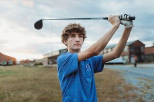 A golfer in a blue shirt practices their swing with a driver against a cloudy sky.