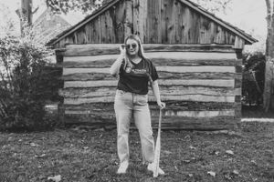 Black and white photo of person in casual attire standing in front of log cabin.