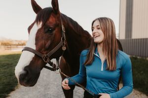 Beautiful bay horse with white blaze standing beside handler outdoors near stable.