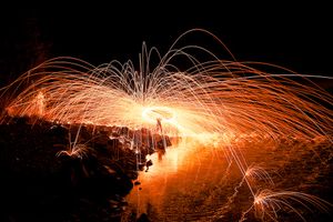 Long exposure photograph of steel wool spinning creating orange light trails reflected on water at night.