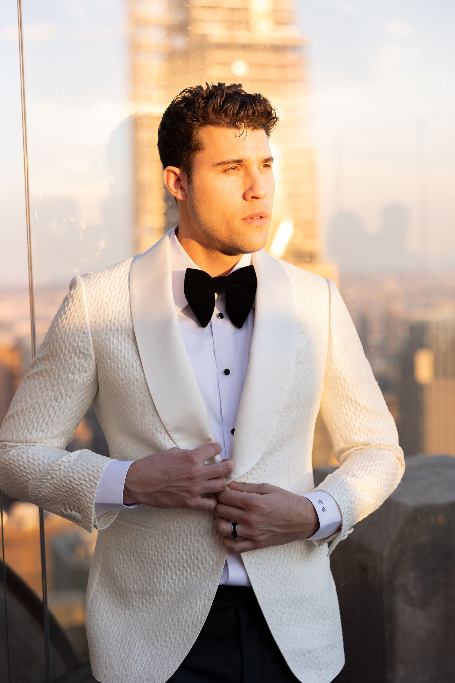 Man in white dinner jacket and bow tie poses against Manhattan sunset backdrop.