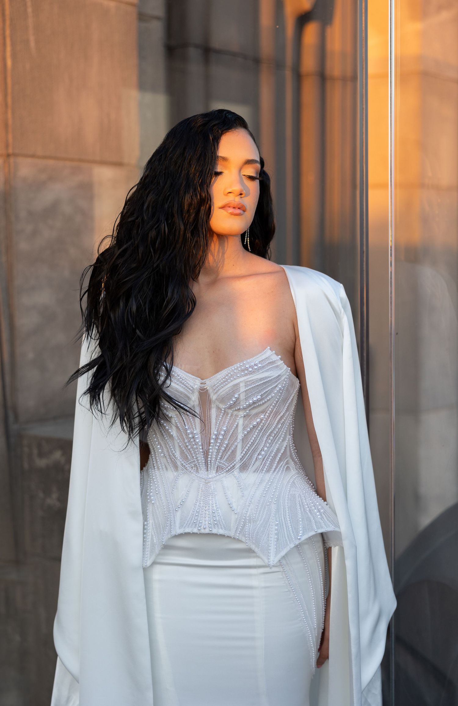 Bride wearing structured white gown and cape in warm golden light.