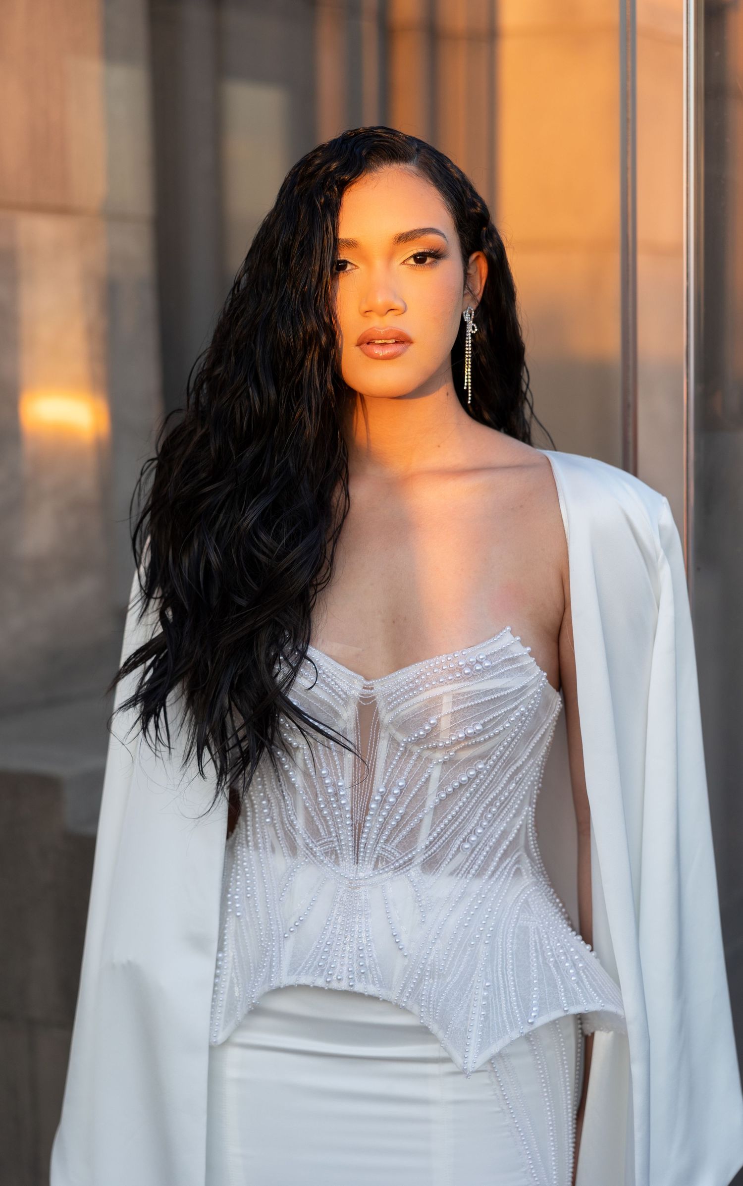 Woman in white corset dress with long dark hair poses elegantly at sunset against city backdrop.