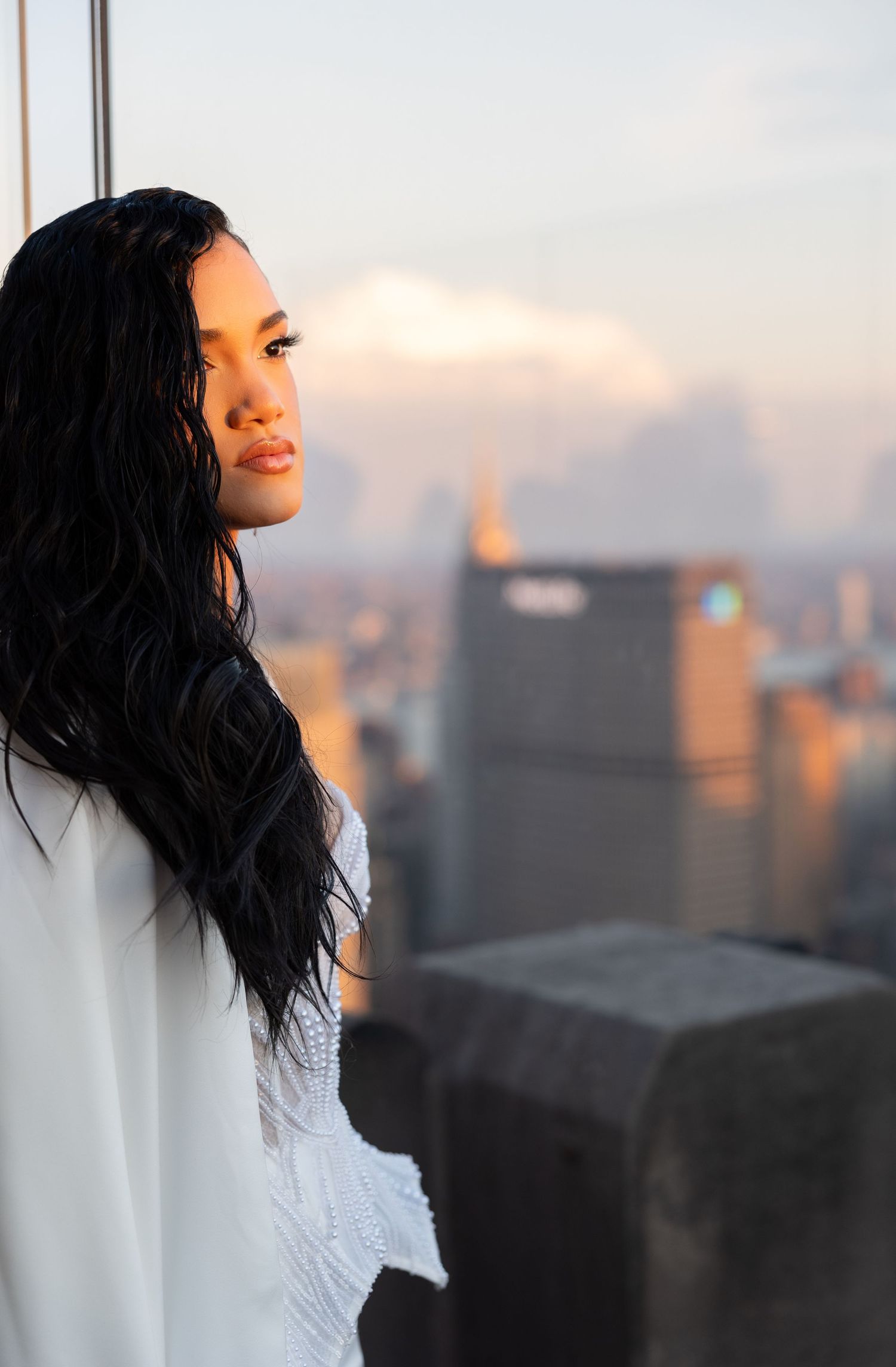 Woman in white dress gazes at sunset city skyline from high-rise perspective.