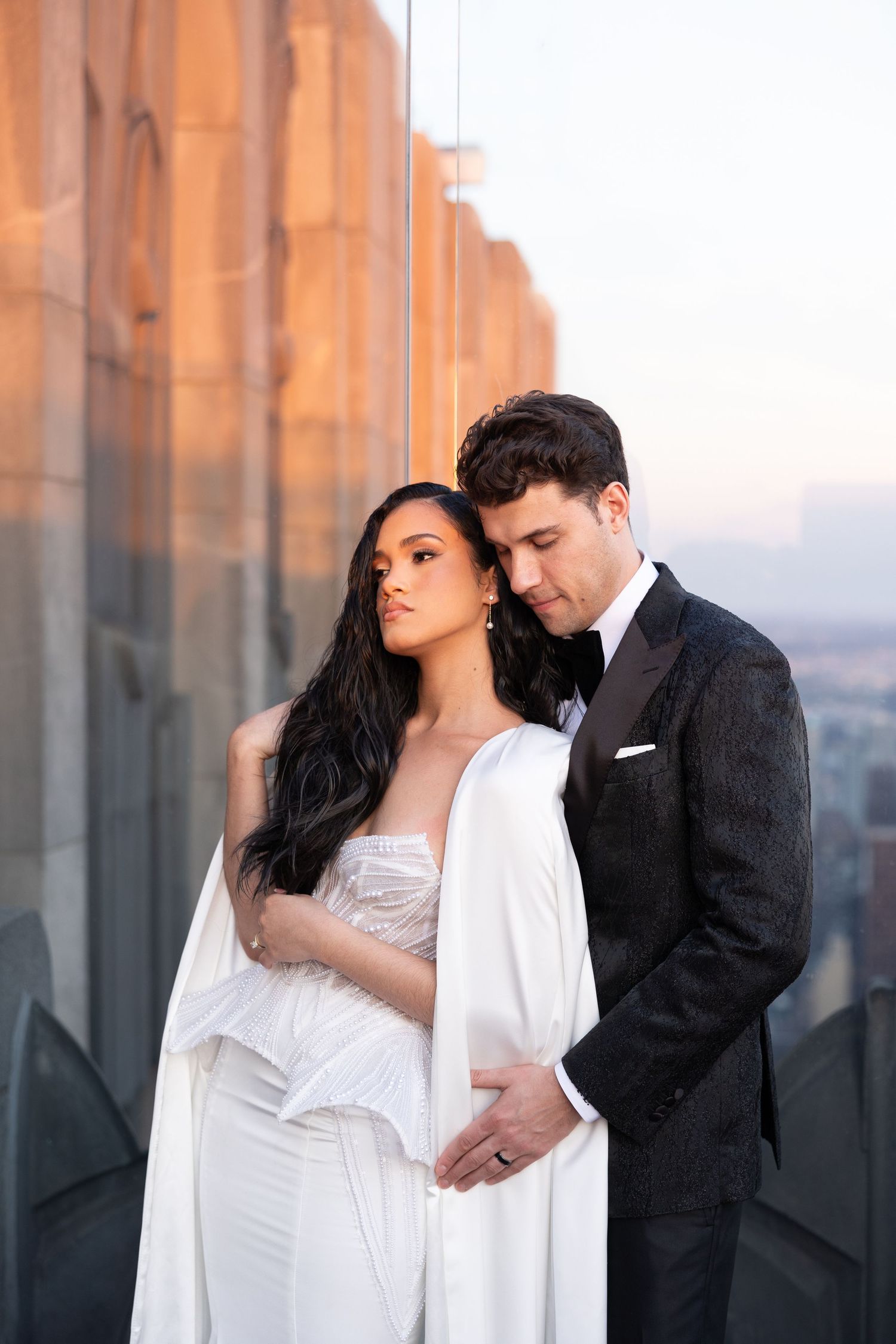 Romantic couple in formal wear embrace against sunset city backdrop.