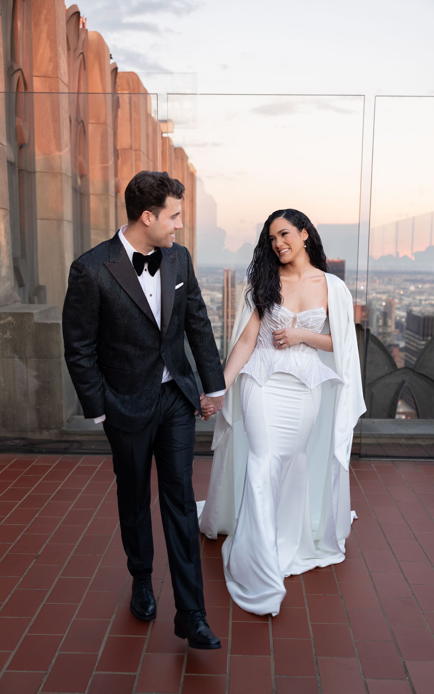 Elegant couple walking hand in hand on a rooftop with city skyline behind them.