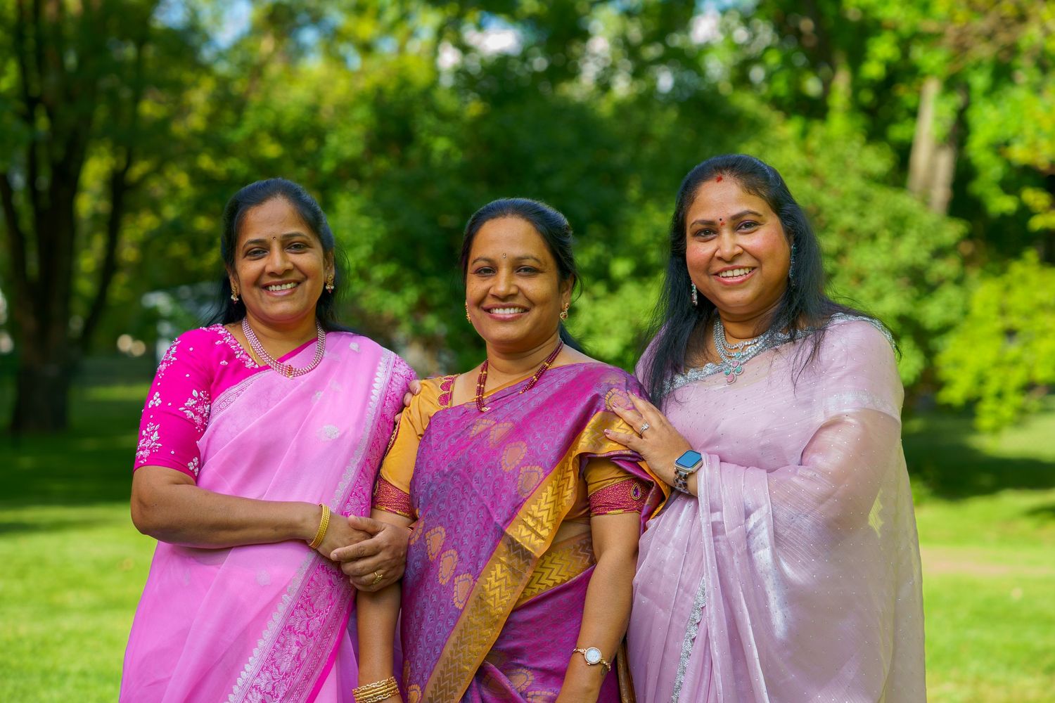 Three women in pink and lavender Indian sarees smiling outdoors in a lush green park setting.