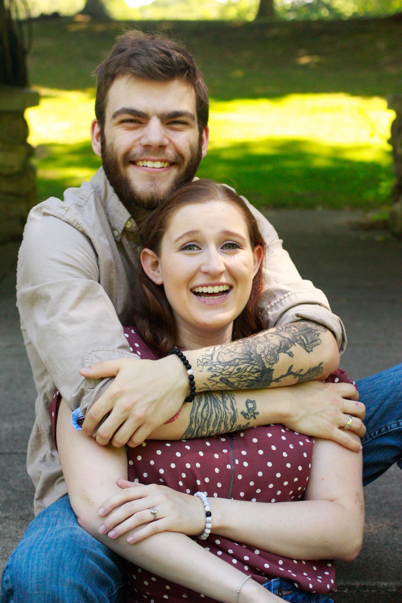 Happy young couple embracing and laughing outdoors in a park, woman has tattoos on her arms.