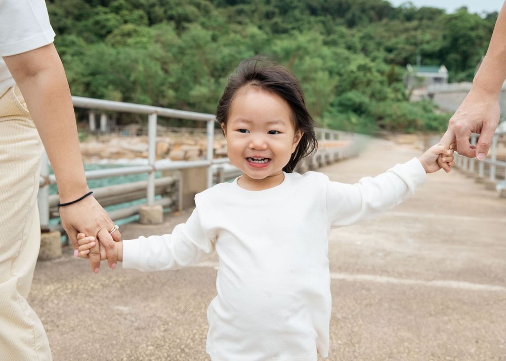A child in white clothing runs joyfully along a sandy path surrounded by greenery on a sunny day.