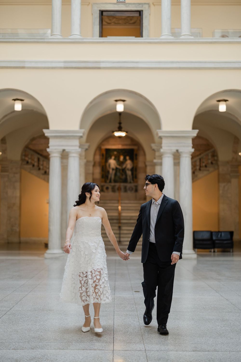 A couple in wedding attire holding hands while walking through an elegant arched hallway with classical architecture.