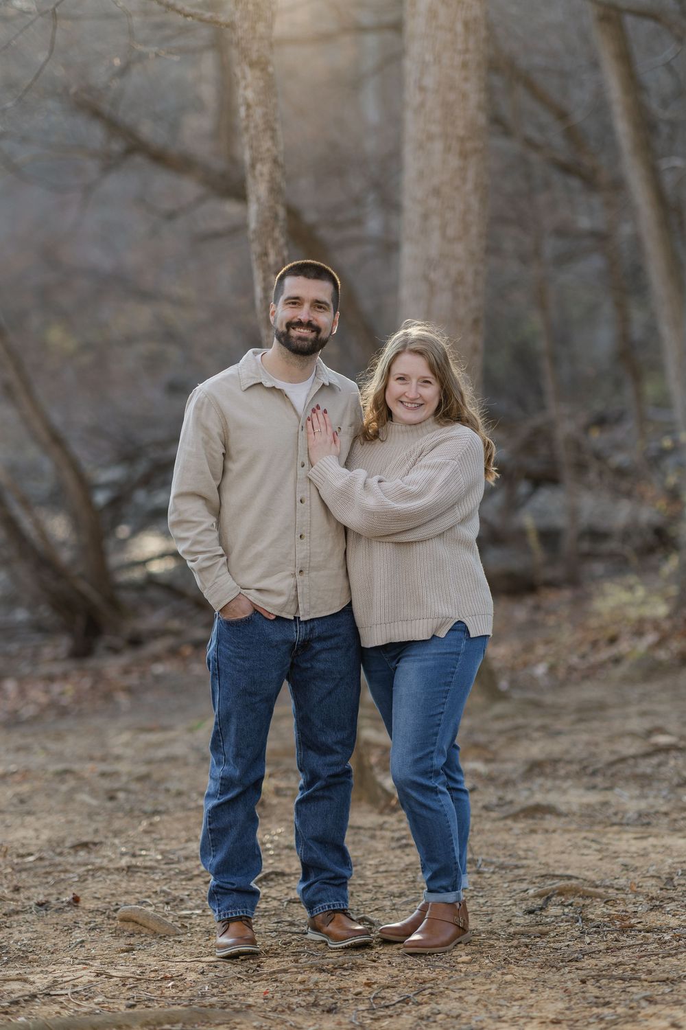 A couple in matching beige sweaters and jeans pose together on a wooded path during an outdoor photo session.