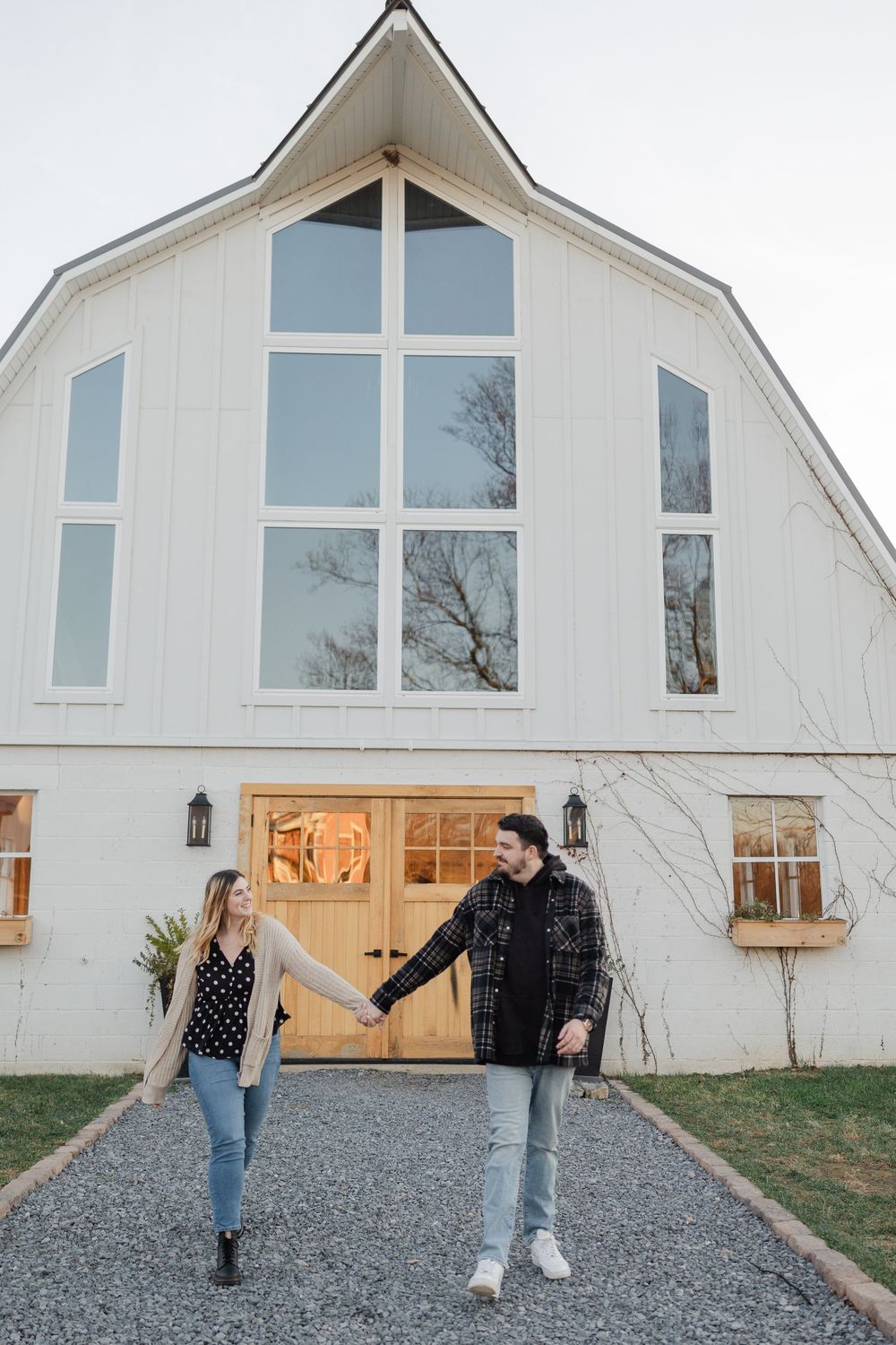 A couple walks together in front of a modern white barn venue with large windows and gravel path.