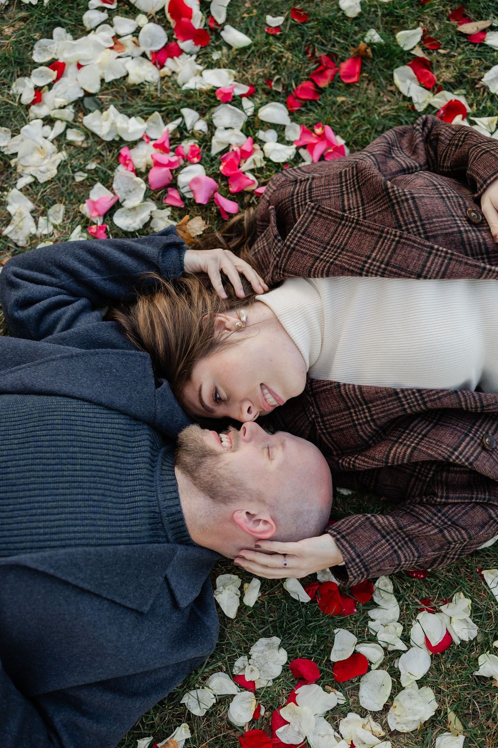 A couple lies on grass surrounded by scattered red and white rose petals in a romantic outdoor setting.