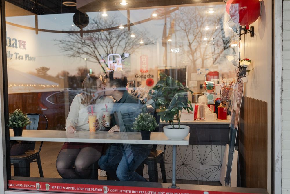 Through a glass window, customers sit at a modern coffee shop counter with warm lighting and reflective surfaces.
