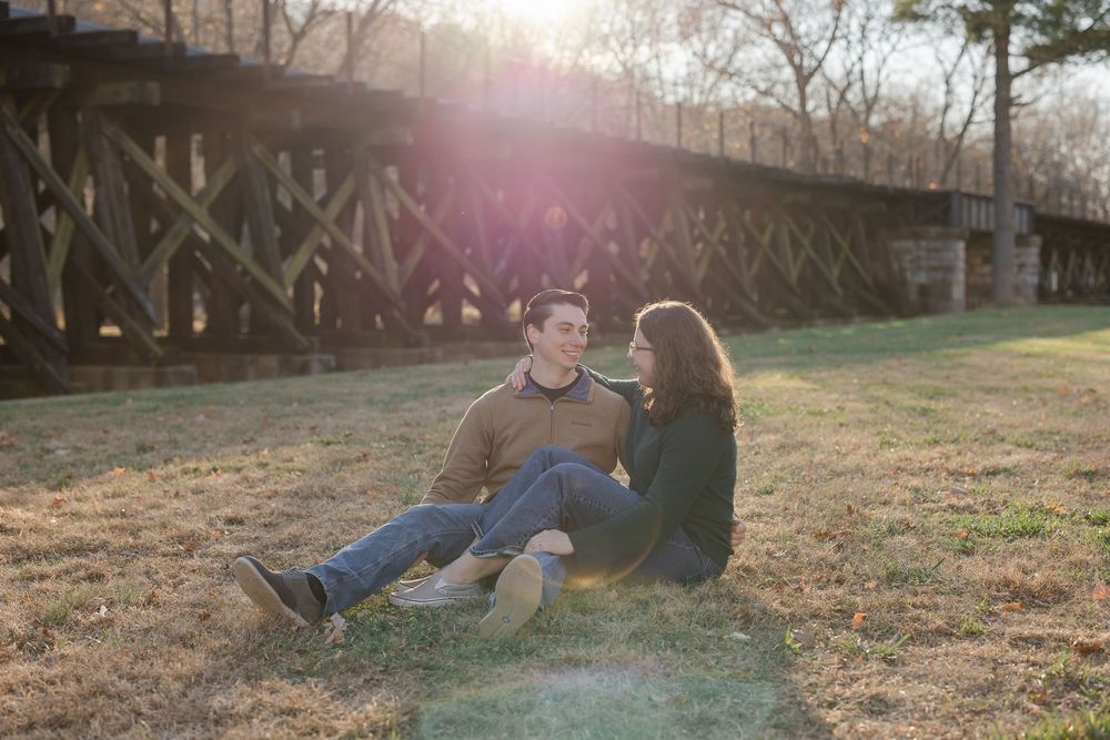 A couple sits together on a grassy hillside during golden hour as sunlight streams through the trees.