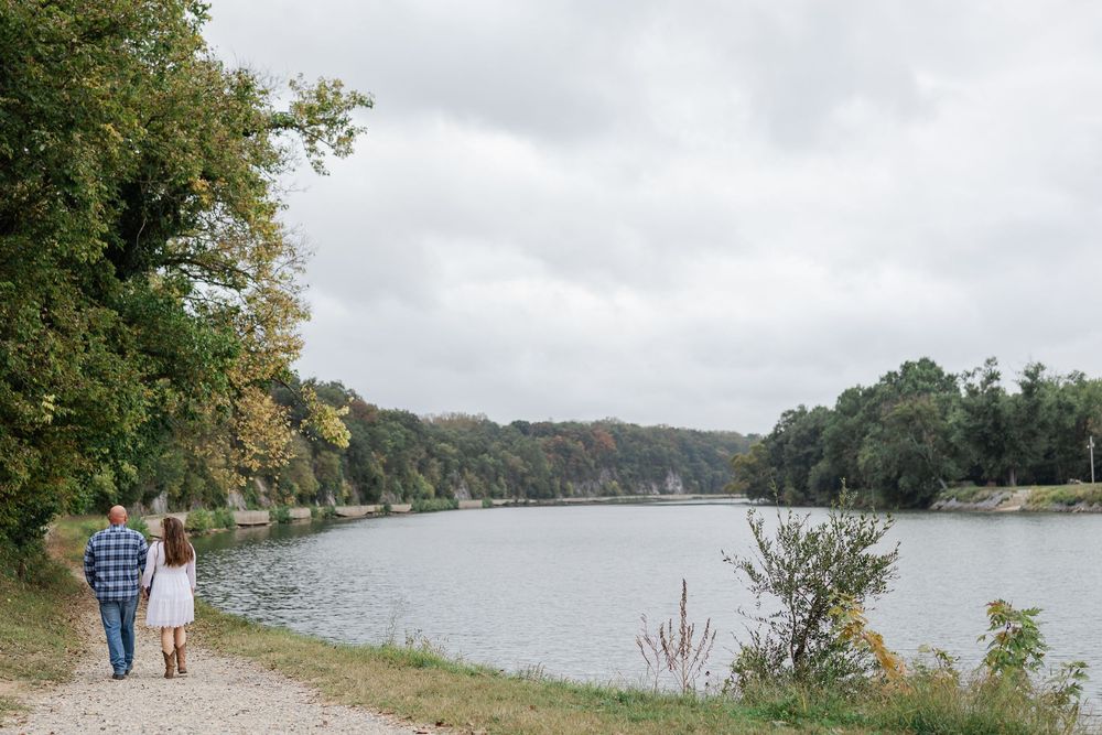 People walk along a scenic lakeside trail surrounded by autumn trees and cloudy skies.