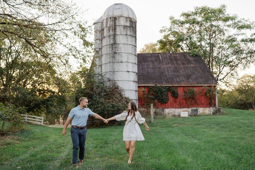 A rustic red barn with a silver silo stands in a lush green field during golden hour.