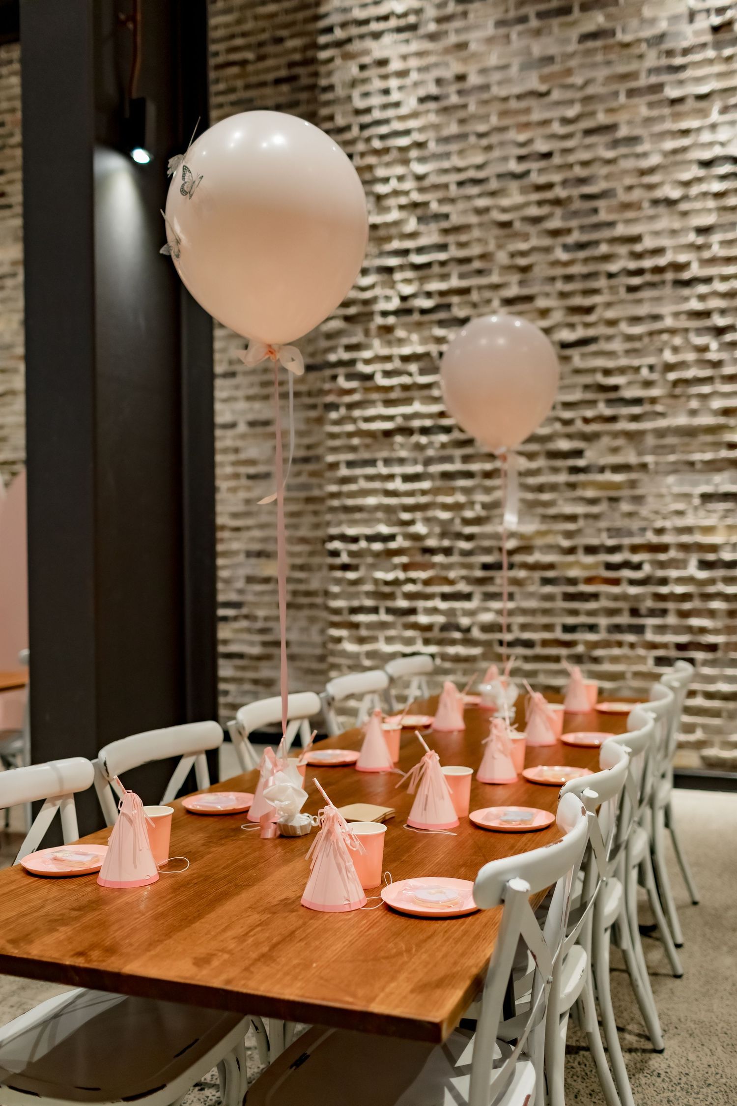 Pink party setup with white balloons and chairs against exposed brick wall for elegant celebration.