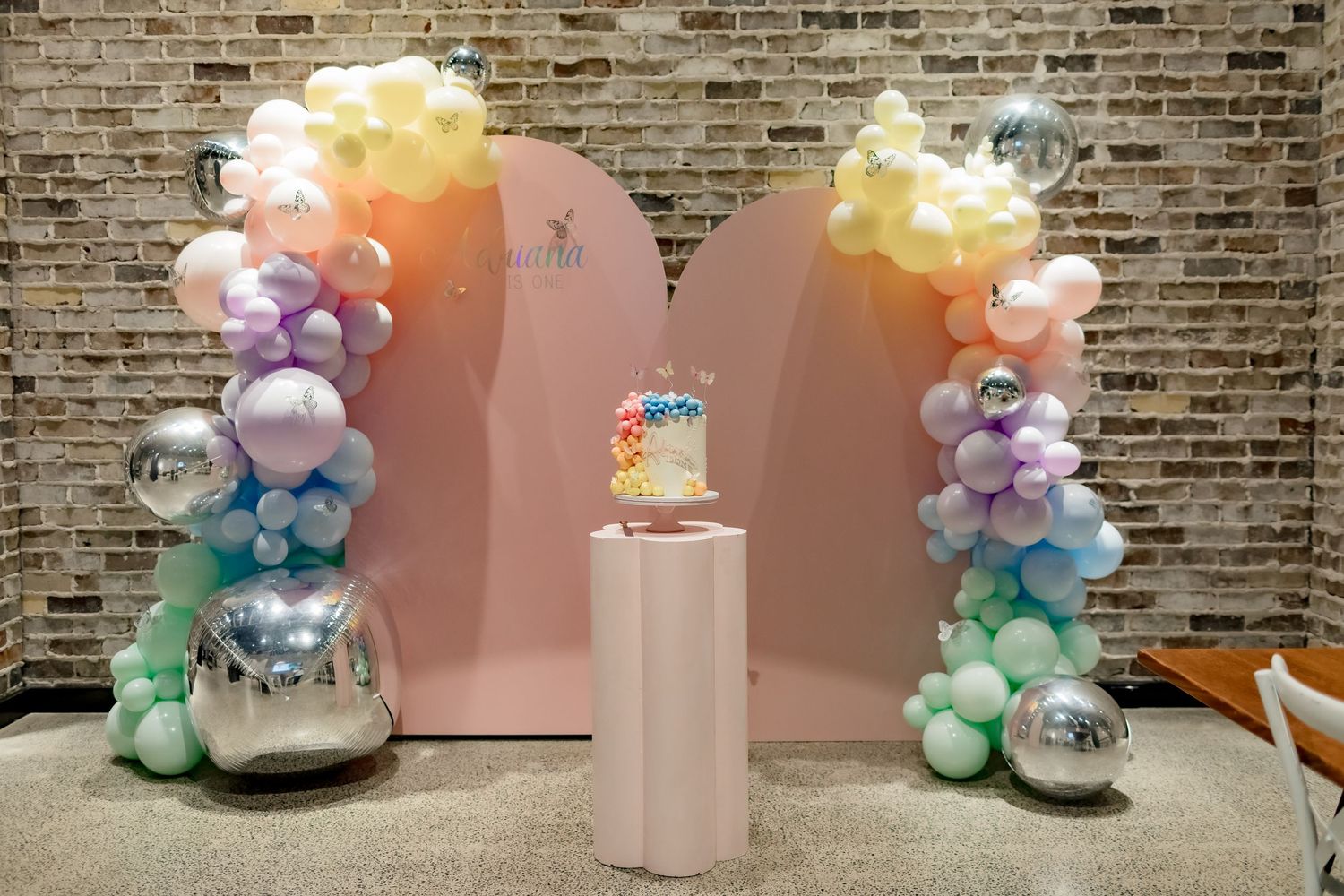 Pastel balloon arch with pink backdrop and decorative pedestal for birthday celebration.