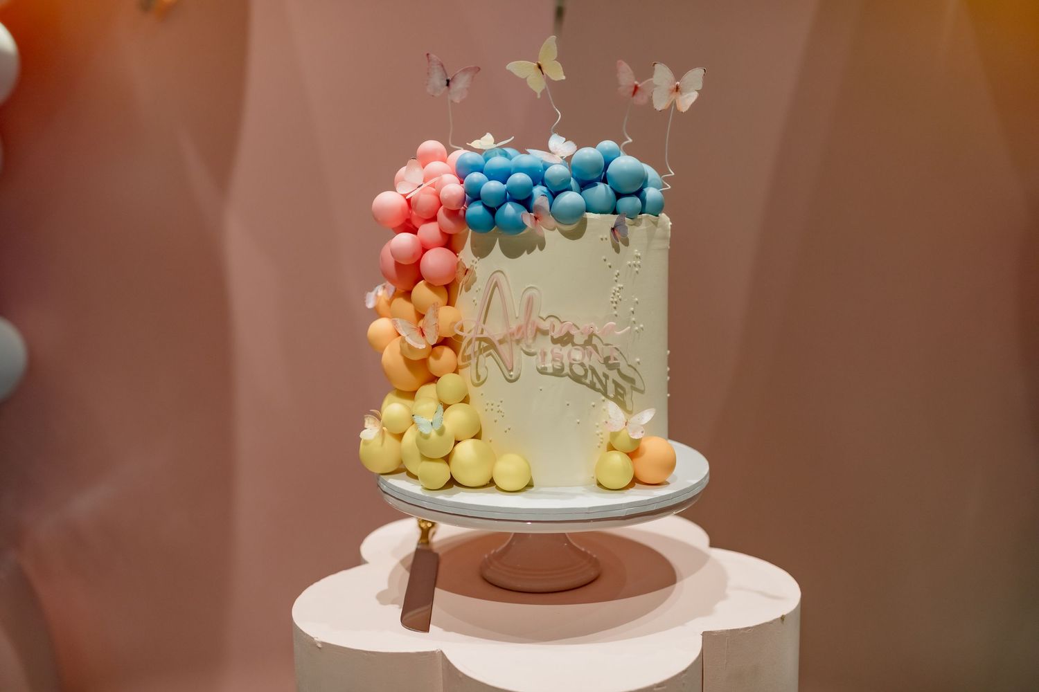 White celebration cake with pastel balloon decorations and butterfly accents on display stand.