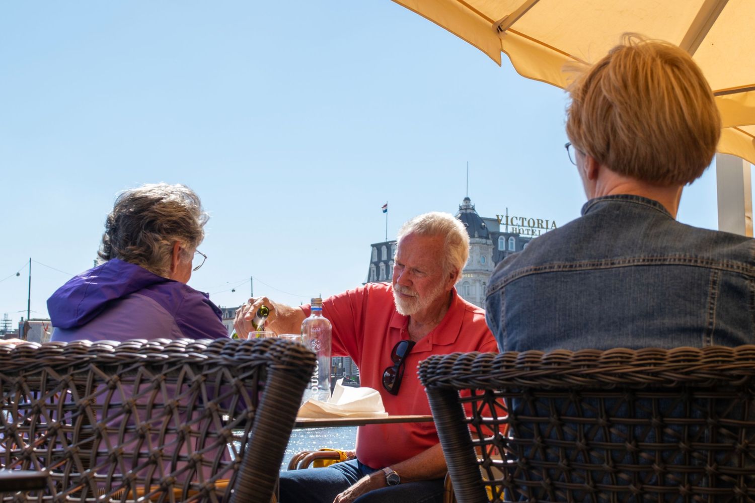 People sitting at outdoor cafe tables with umbrellas near harbor.
