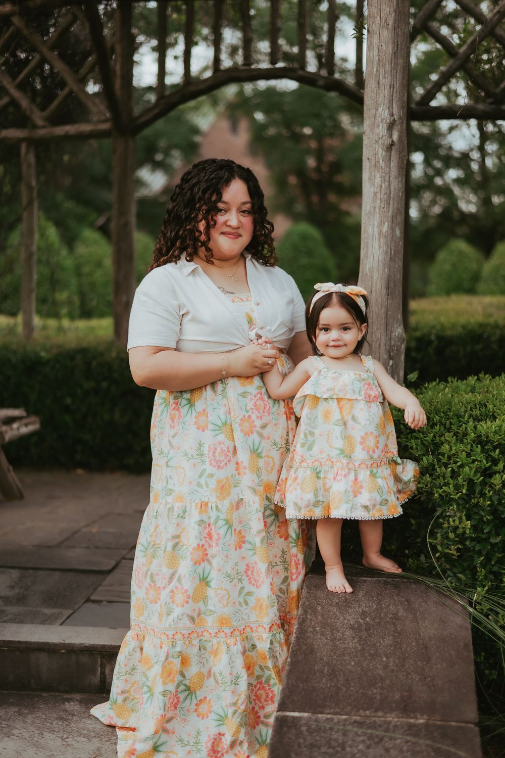 A mom and her young daughter wearing matching floral dresses pose together on wooden steps outdoors.