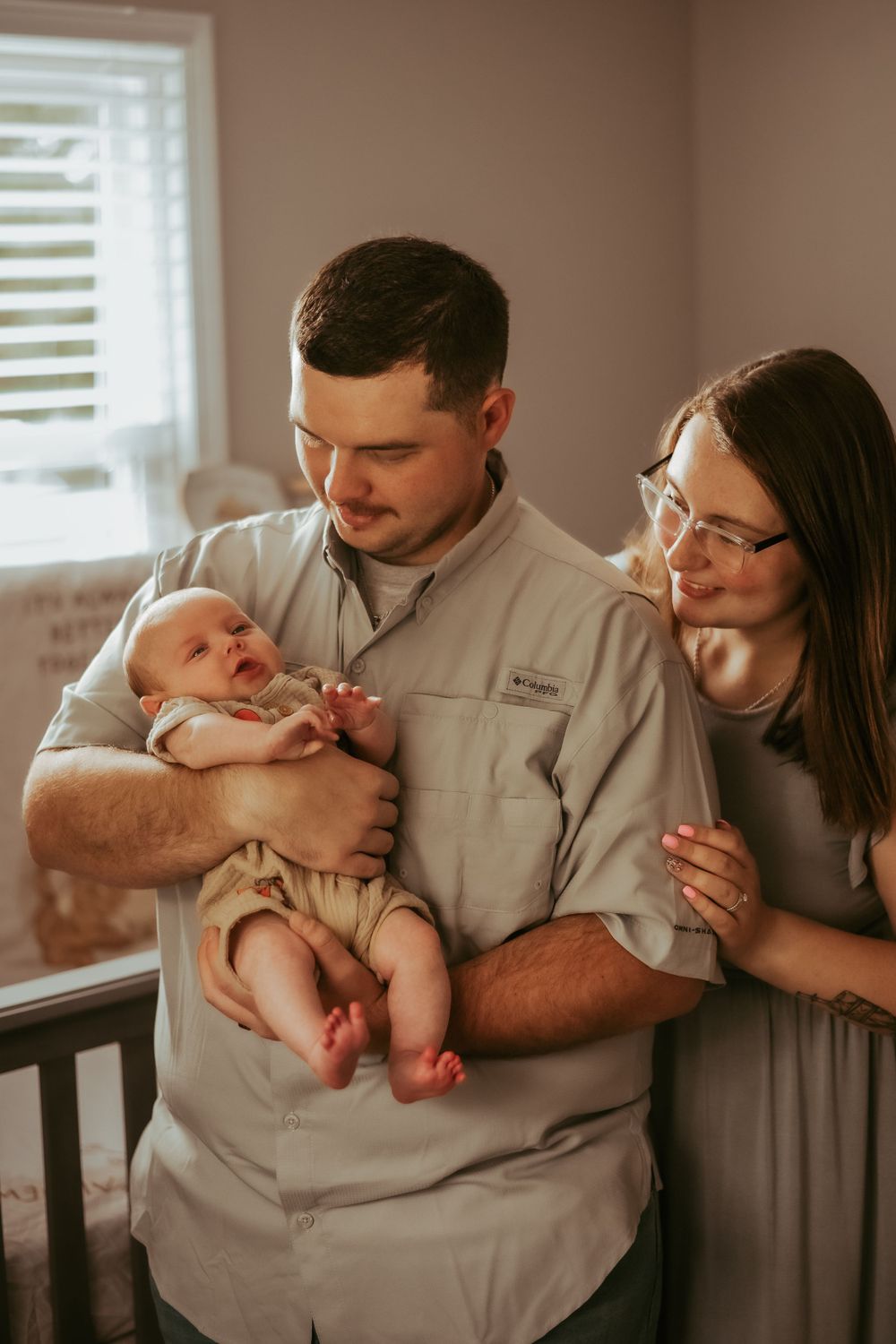 New parents smile while holding their infant in nursery with window light.
