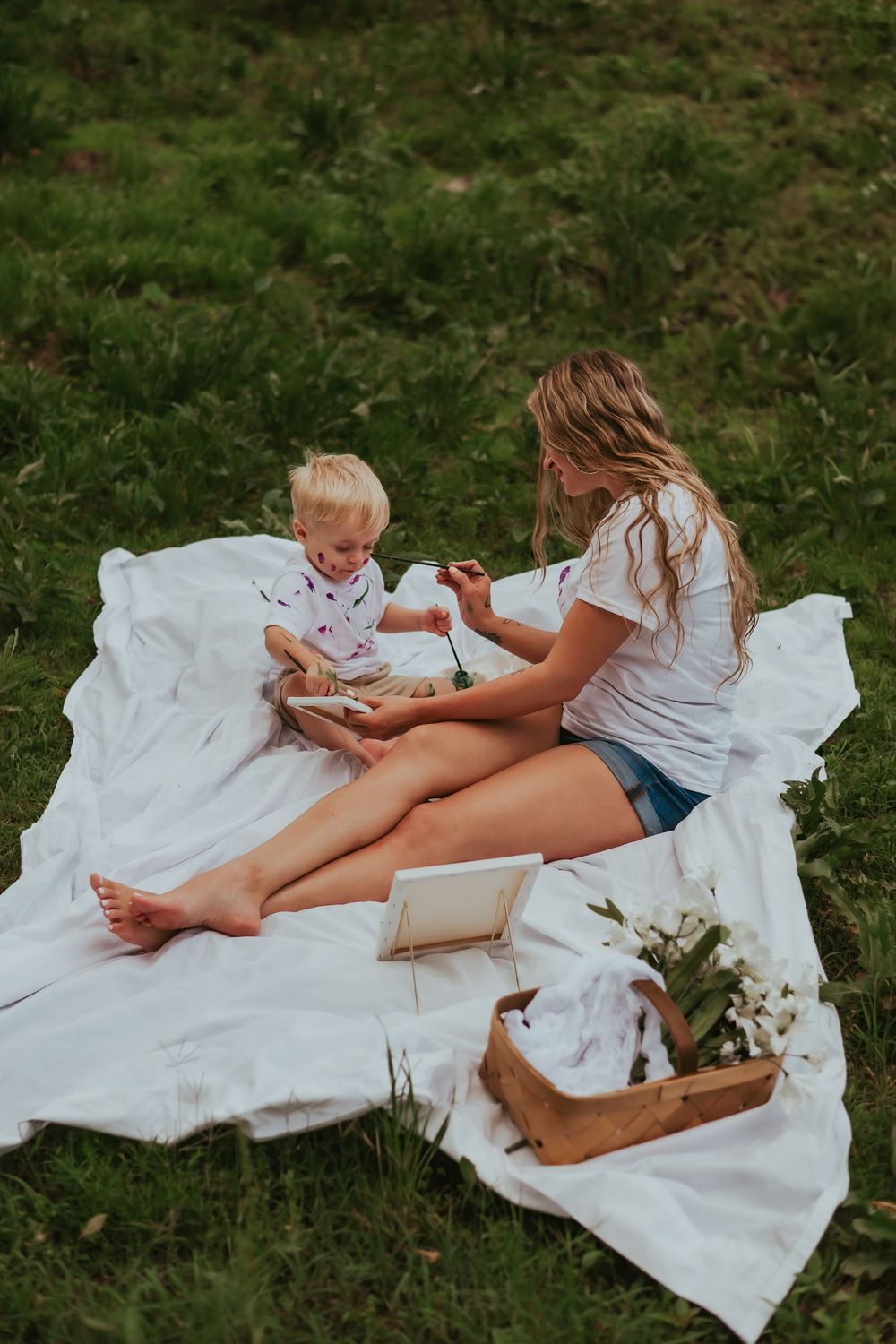 A mom and her young son paint canvases on a white blanket outdoors in lush green grass.