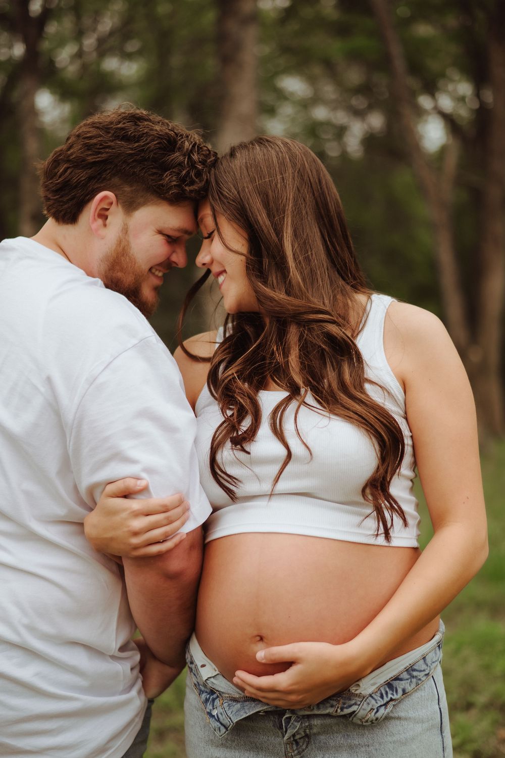 Intimate maternity photo shows couple in white tops embracing tenderly in wooded outdoor setting.