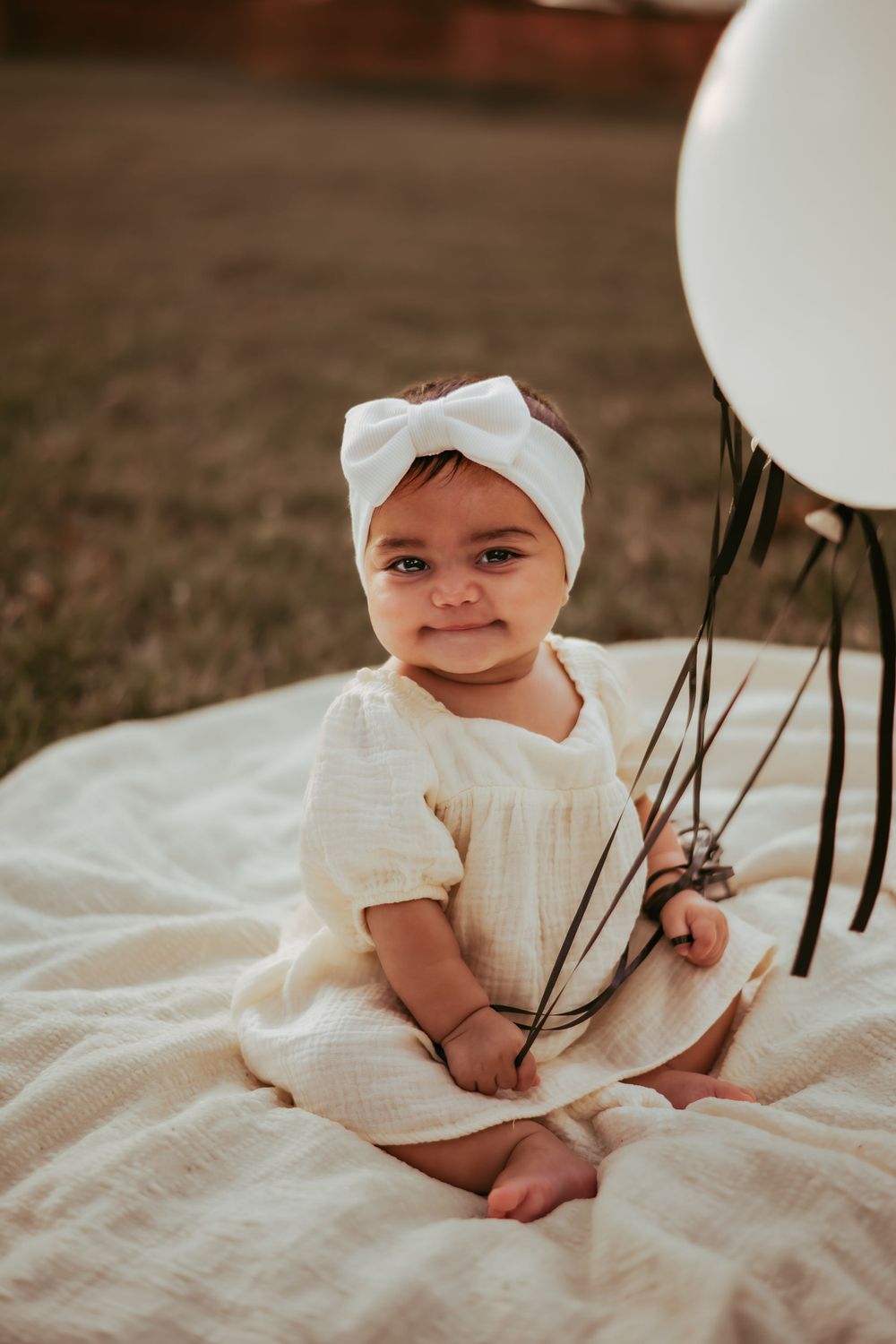 Baby in white dress and bow headband sits on blanket holding black balloons on brown grass.