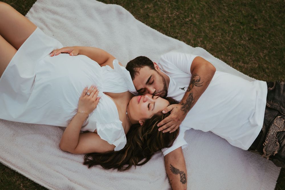 A couple lay on a white blanket in the lush green grass. The man caresses the woman's head that is laying on his shoulder.