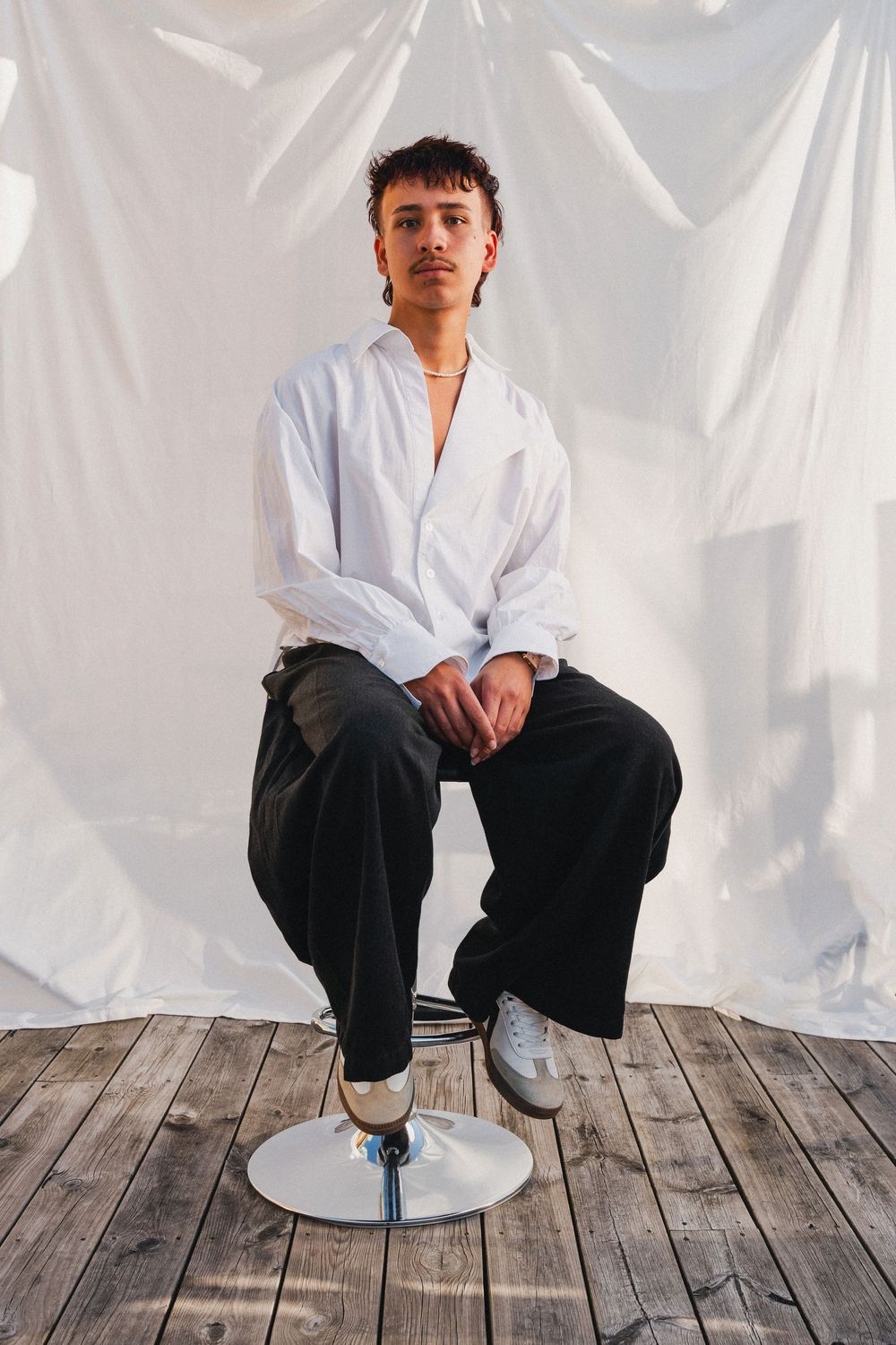 Porträtt av person i vit skjorta och svarta byxor sittande på pall mot ljus tygbakgrund och trägolv. Fotograf Gabriel Alenius