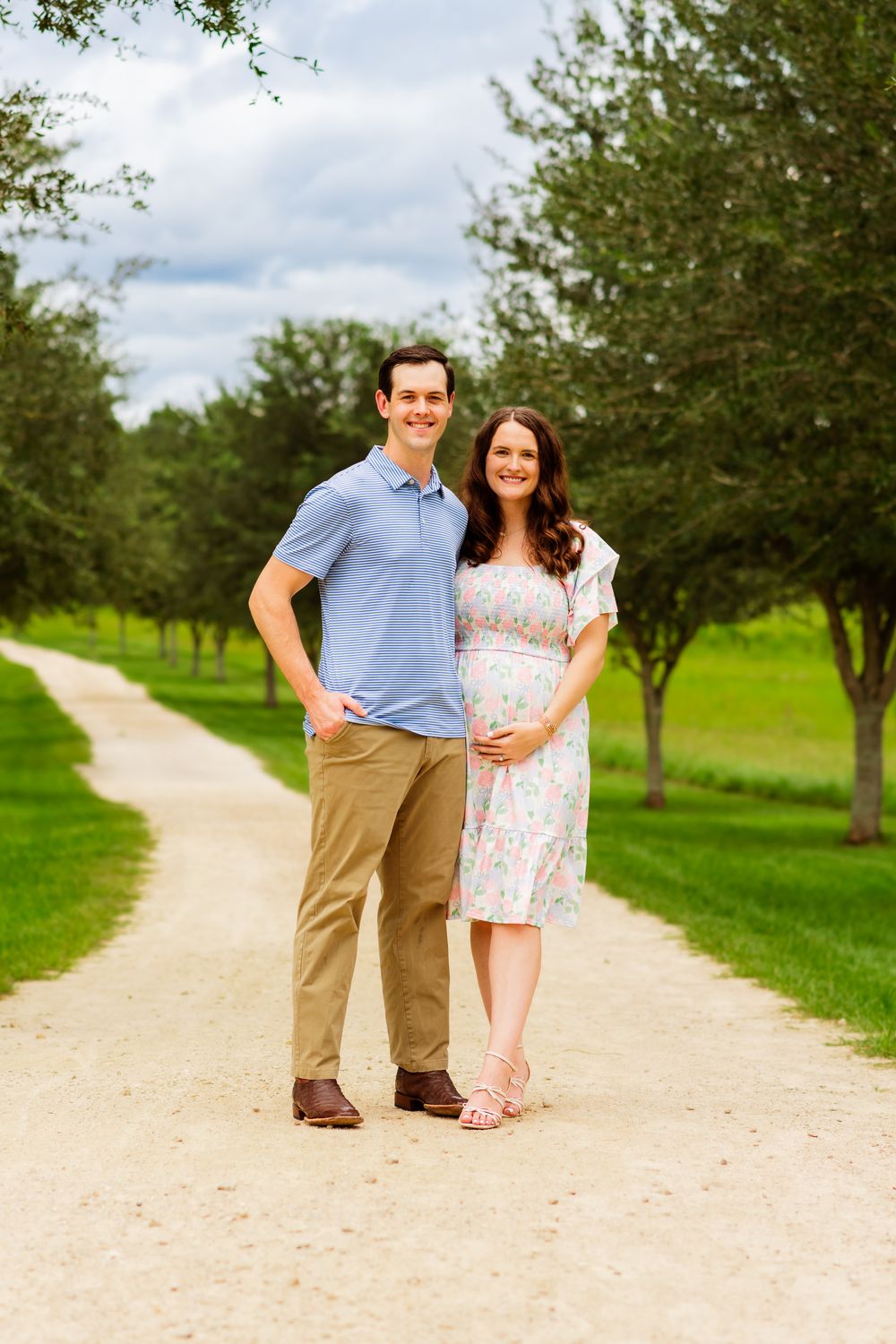 Couple poses on a tree-lined path, woman in floral dress cradling baby bump, man in blue polo and khakis.