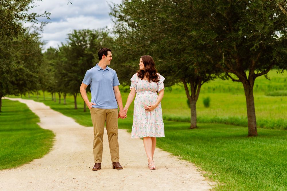 Pregnant couple holding hands on a scenic gravel path surrounded by lush green trees and grass.