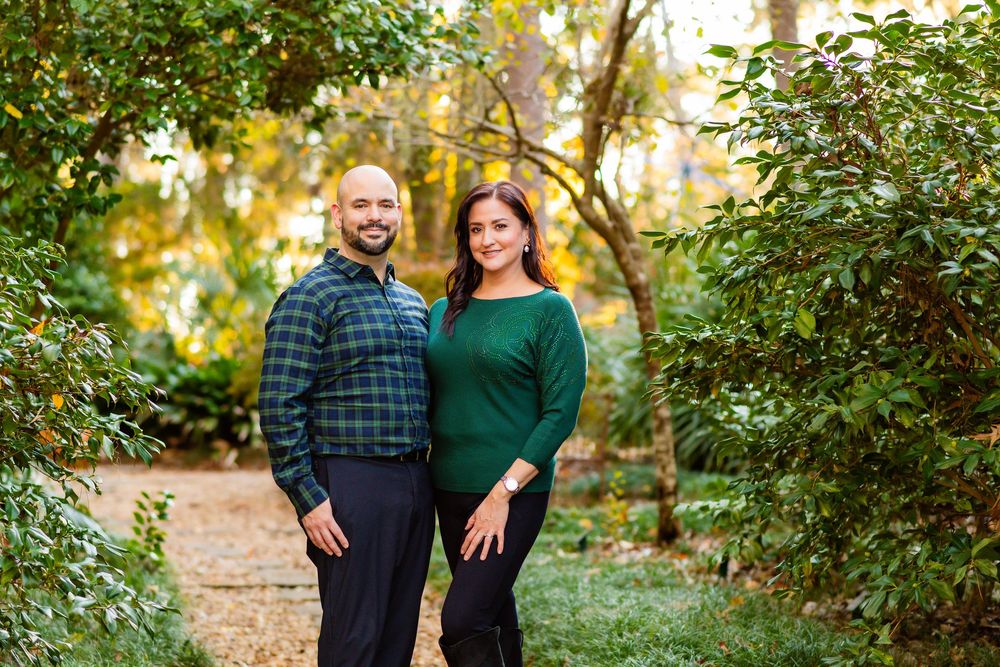 A couple stands together on a leaf-covered path in a lush green garden during golden hour.