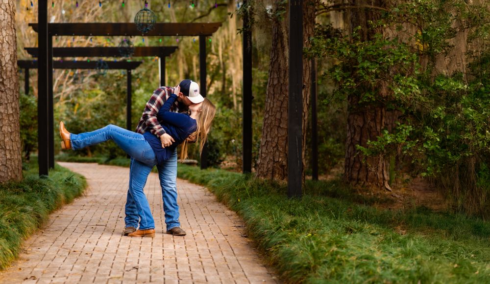 A couple embraces while kissing on a brick path surrounded by wooden pergola and green forest scenery.