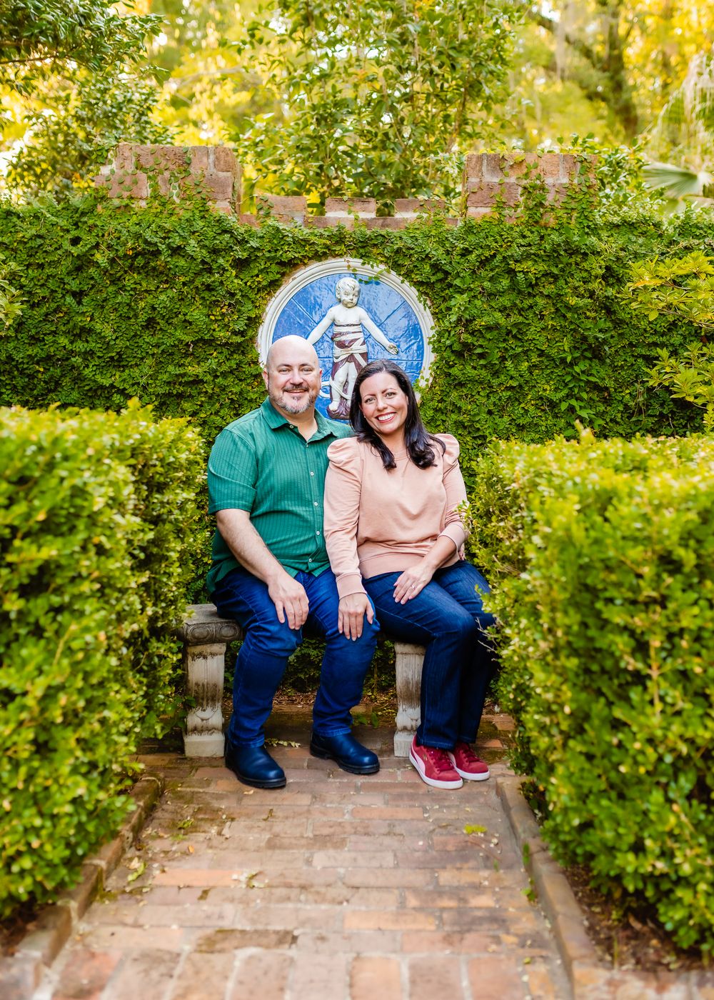 A couple sits together on brick steps surrounded by lush green hedges in a garden setting.