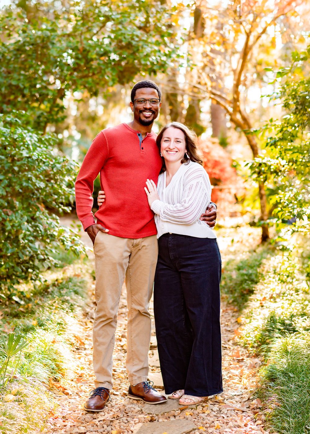 A couple poses together on a sunlit autumn trail surrounded by fallen leaves and trees.