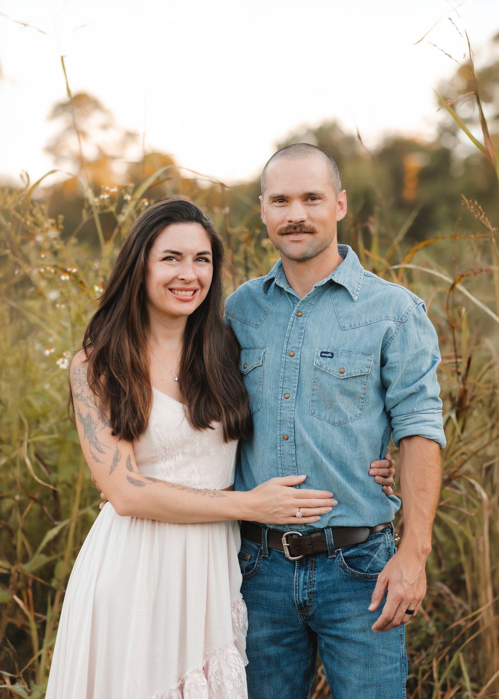A couple poses together in a golden field at sunset wearing casual denim and white clothing.