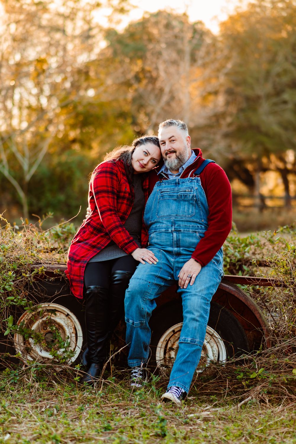 Couple in red flannel and denim overalls posing on rustic farm equipment at sunset.