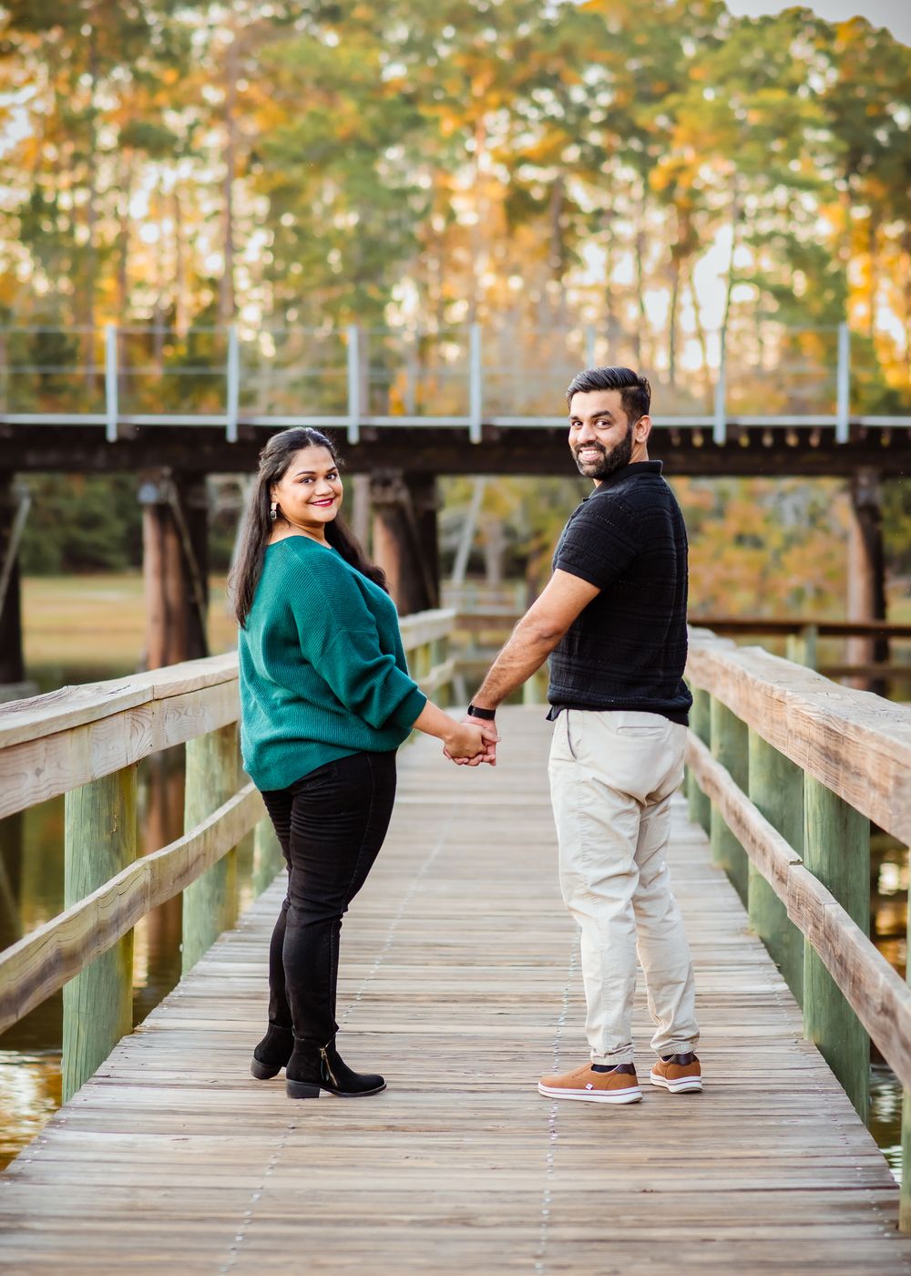 Couple walking and posing playfully on a wooden boardwalk surrounded by autumn trees at sunset.