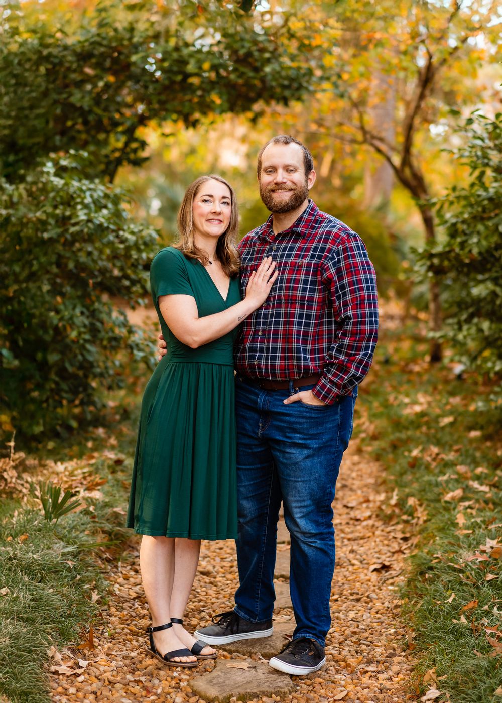 A couple poses together on a leaf-covered autumn path in a forest wearing a green dress and plaid shirt.